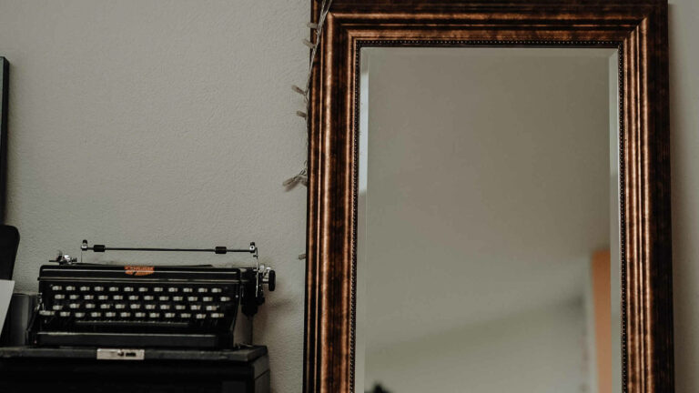 Figurenbeschreibung - per Spiegelszene? Eine alte schwarze Schreibmaschine steht neben einem altmodischen Spiegel mit dickem Goldrand vor einer hellen Wand. Photo by Laura Chouette via Unsplash.